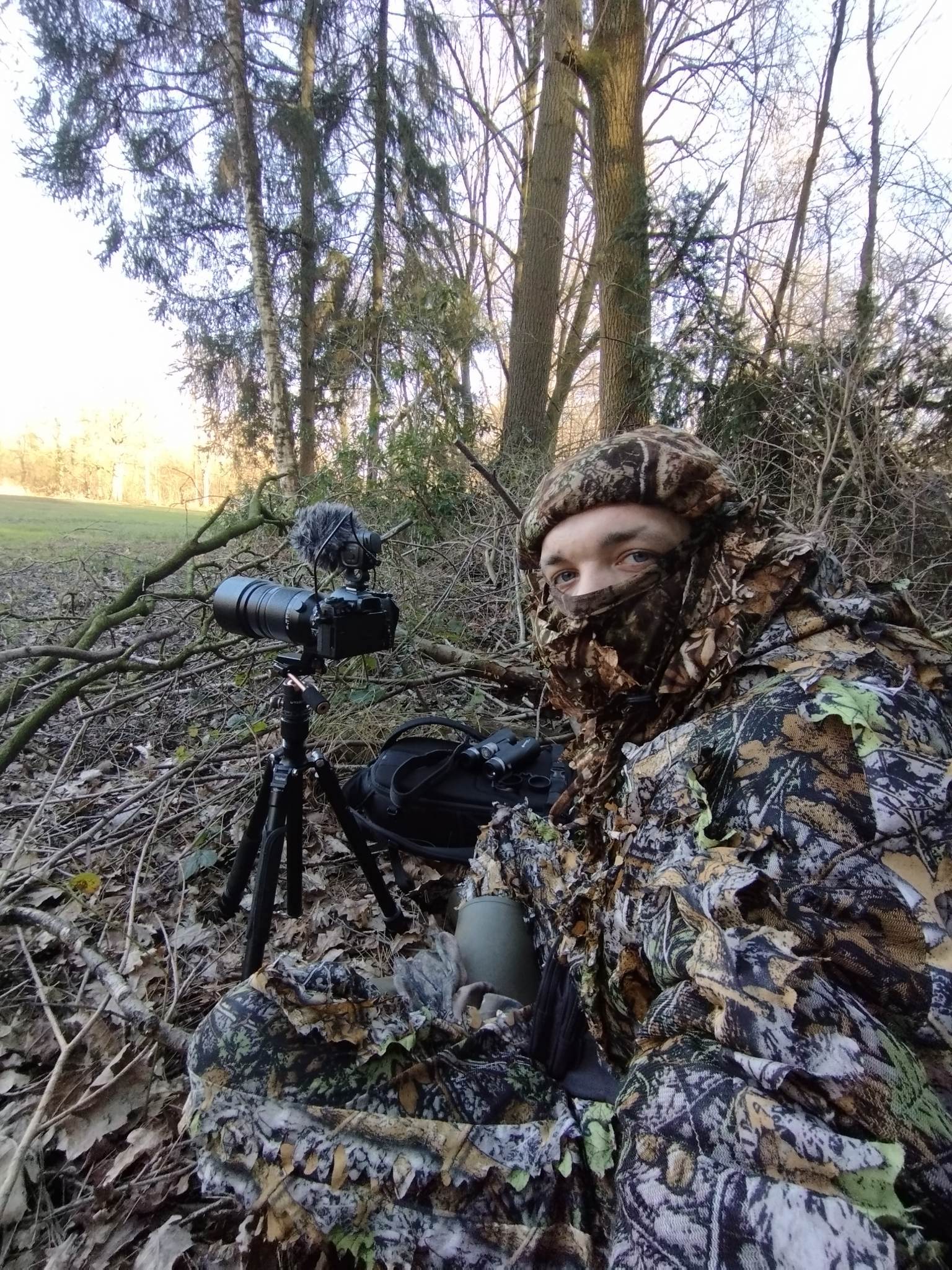 Arne Moons, photographer – man in a camo suit lying down among dried leaves next to a tripod with a camera mounted on it and some biloculars.