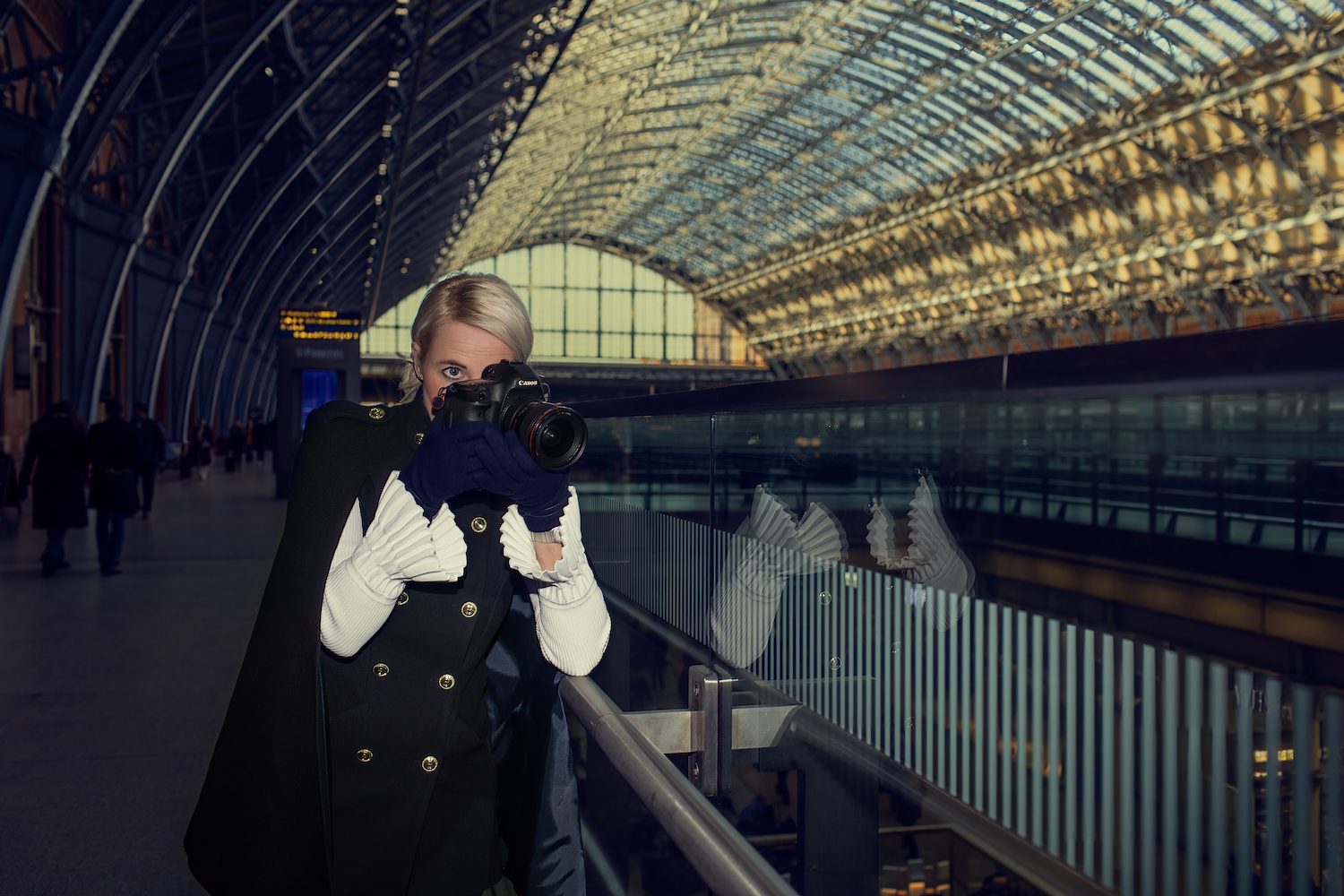 Herma Kluin – blond elegant woman wearing a blue coat and white blouse, taking pictures of someone while inside a train station