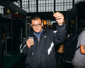 Ein junger Mann hält einen Joint in die Kamera, im Hintergrund sieht man Stände