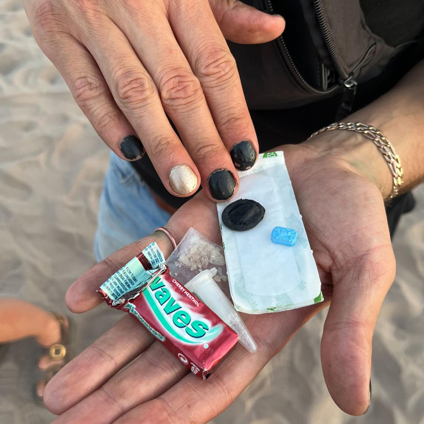 In der Hand sind drei verschiedene chemische Drogen, ein Fingerkondom, ein Pflaster und Kaugummis. Der Mann hat schwarz lackierte Fingernägel und trägt ein goldenes Armband.