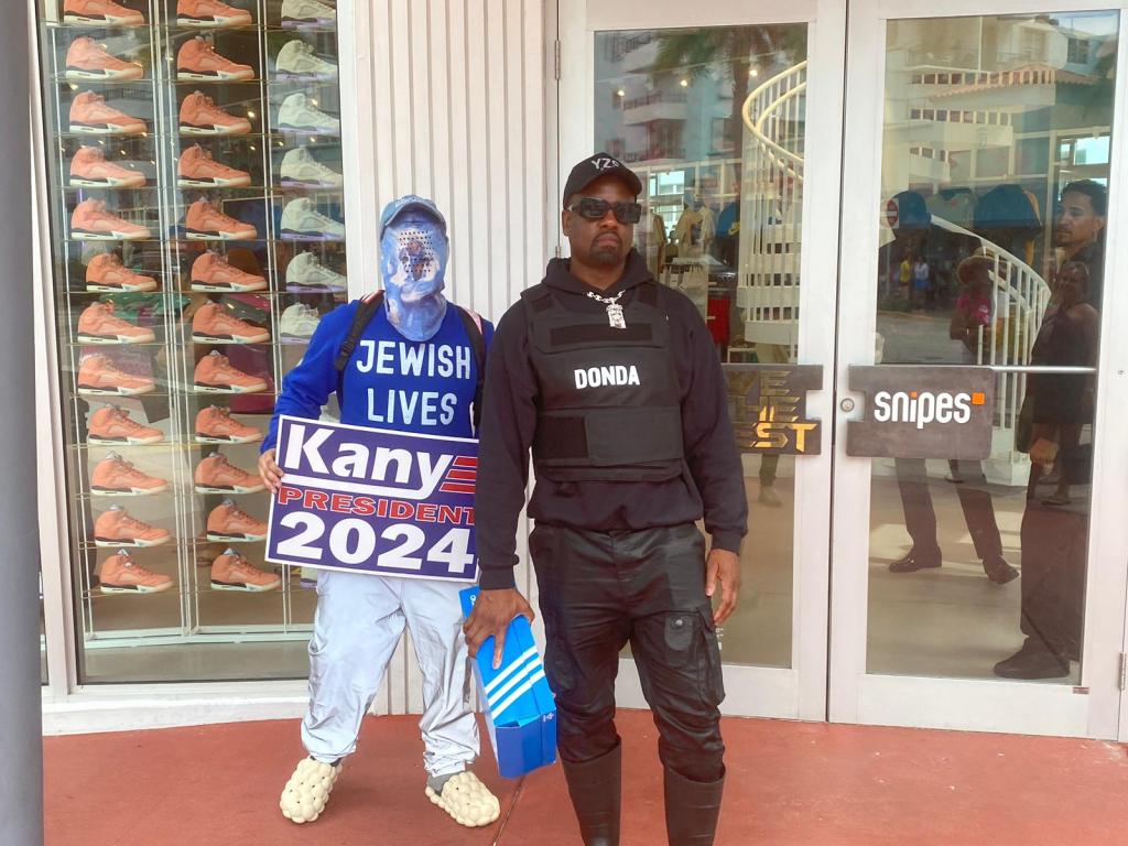 Kanye West impersonator holding a shoe box with a person holding a 'Kanye 2024' sign