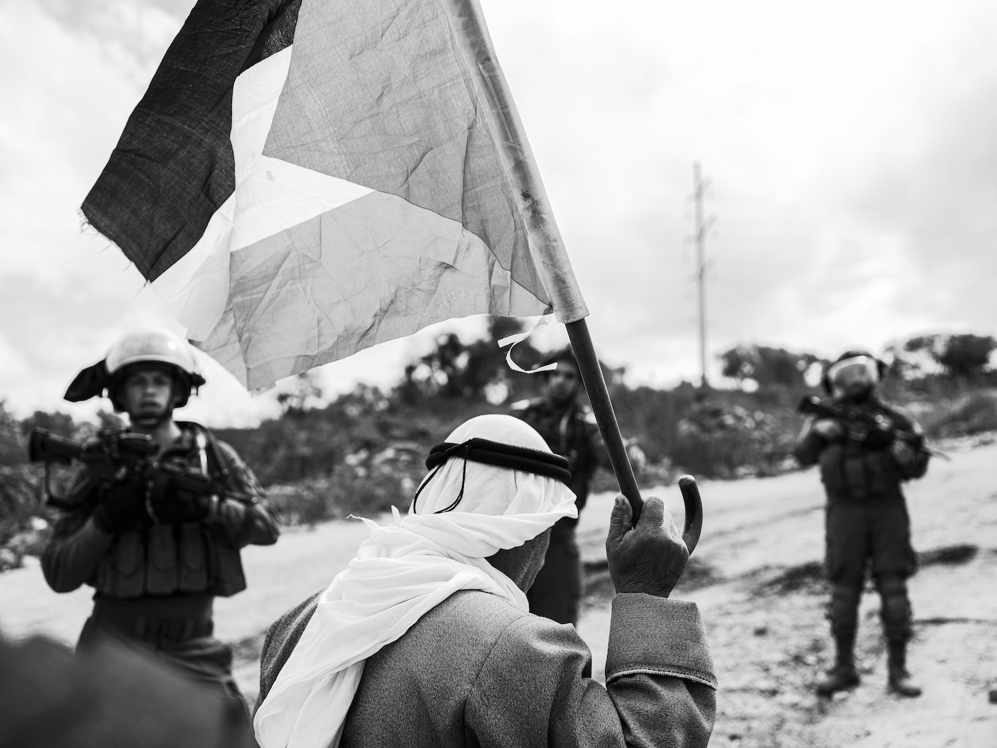 An old man waves a Palestine flag in front of three Israeli soldiers holding rifles.