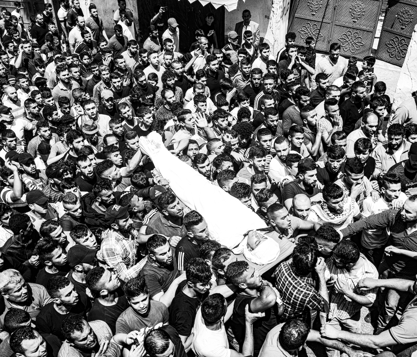 A body wrapped in white cloth lying on a stretcher is carried by a crowd of hundreds of people.