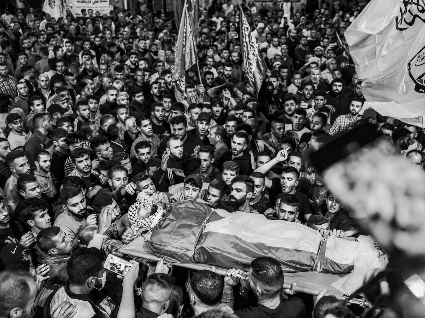 A body wearing a keffiyeh wrapped and wrapped in a Palestinian flag is carried by a crowd.