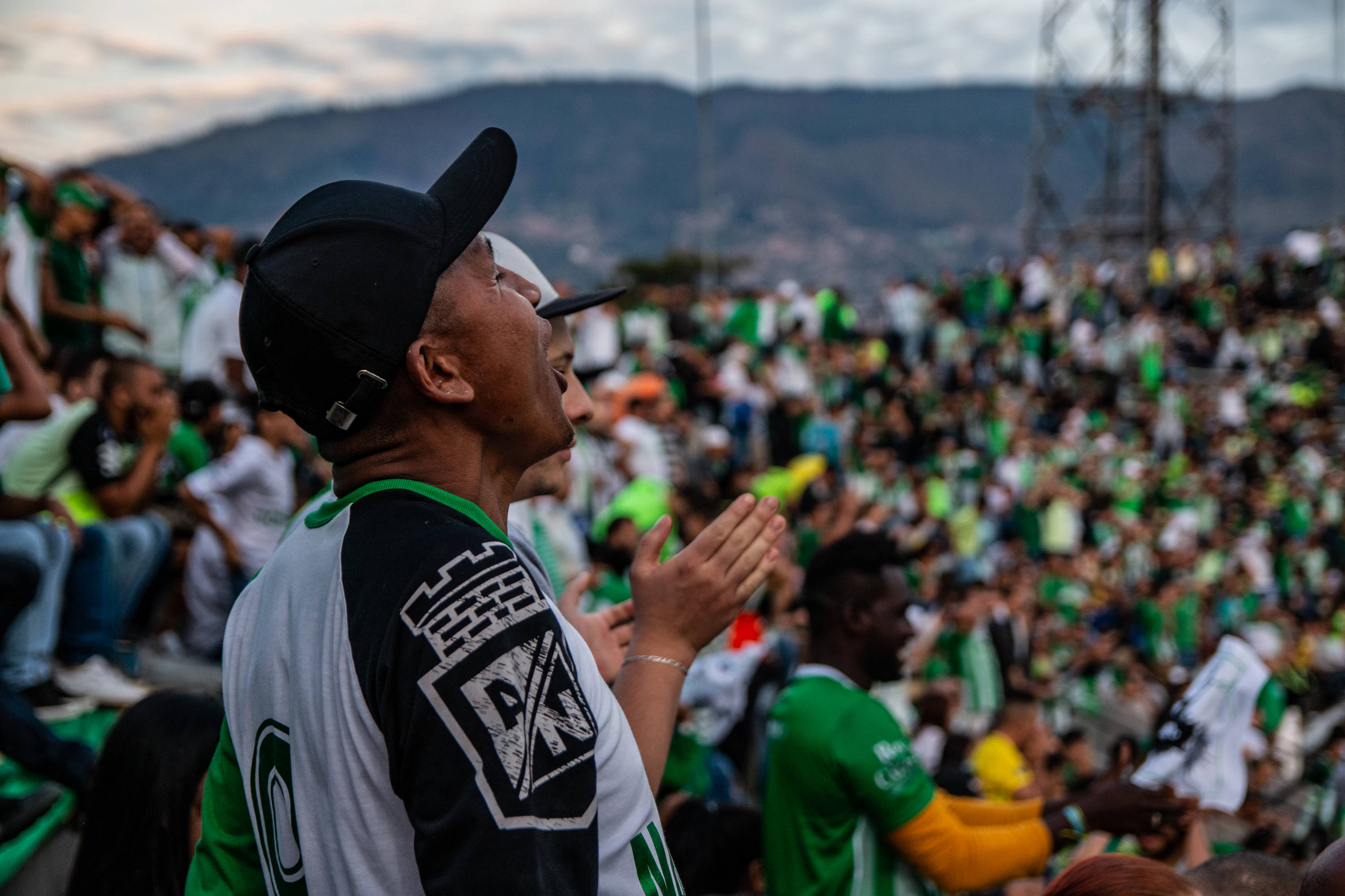 A man wearing a black, white and green football shirt and a black cap screams in a stadium full of supporters wearing the same shirt.