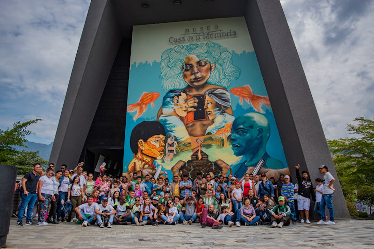 A big group of kids and adults standing in front of a building covered with a colourful fresco.