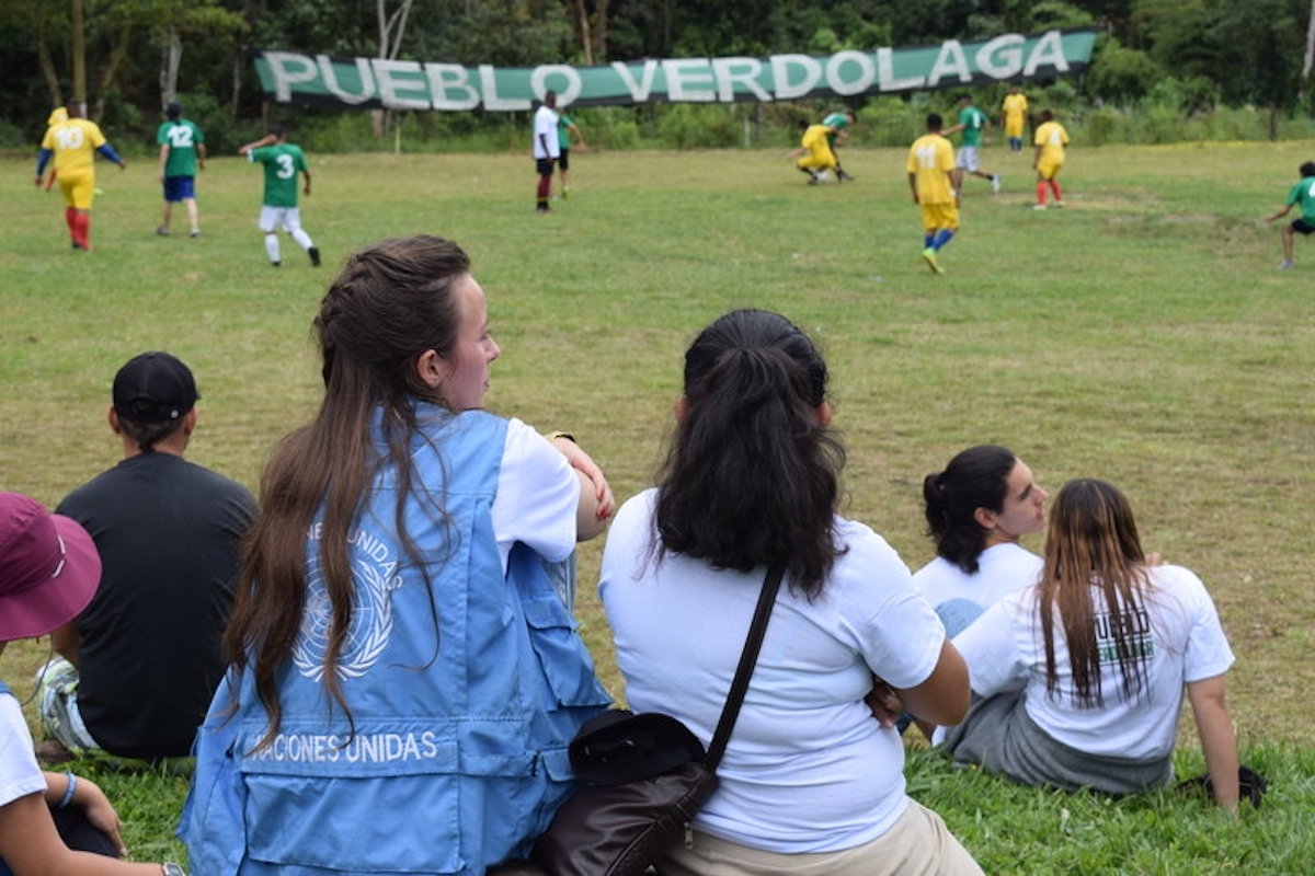 Adults watching teenagers play football at a field.