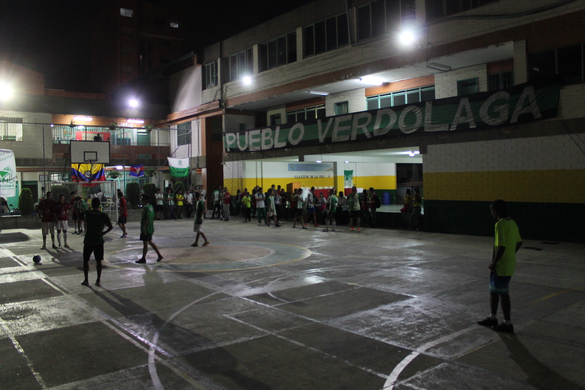 A football and basketball court with teenagers playing in the dark, as a huge flag with Pueblo Verdolaga written on it is visible in the background.