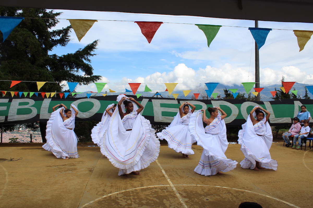 Girls dressed in white traditional dresses dancing on a footfall field with colourful garlands and a flag.