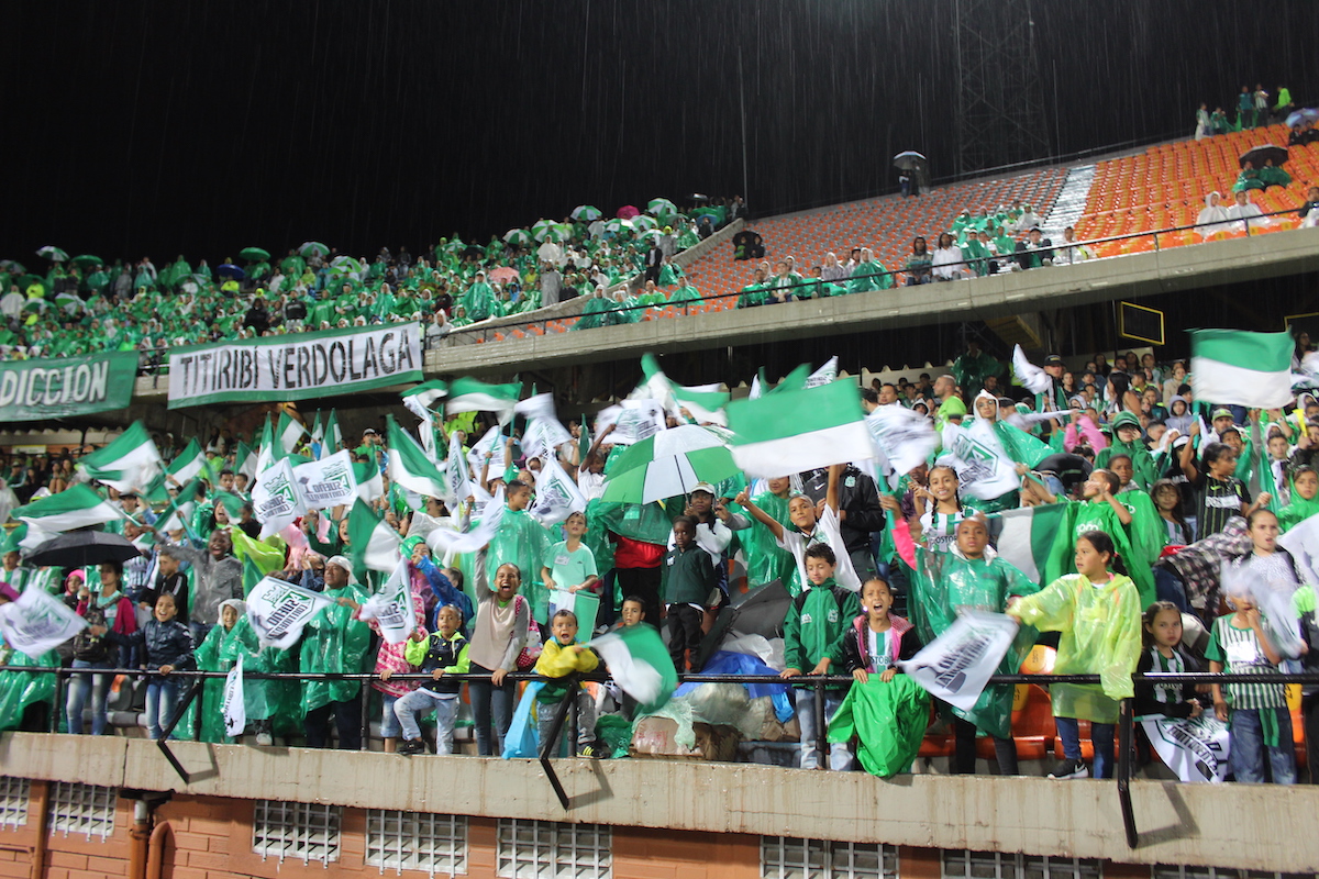 Football supporters dressed in green and while shirts wave flags, banners and umbrellas in a stadium.