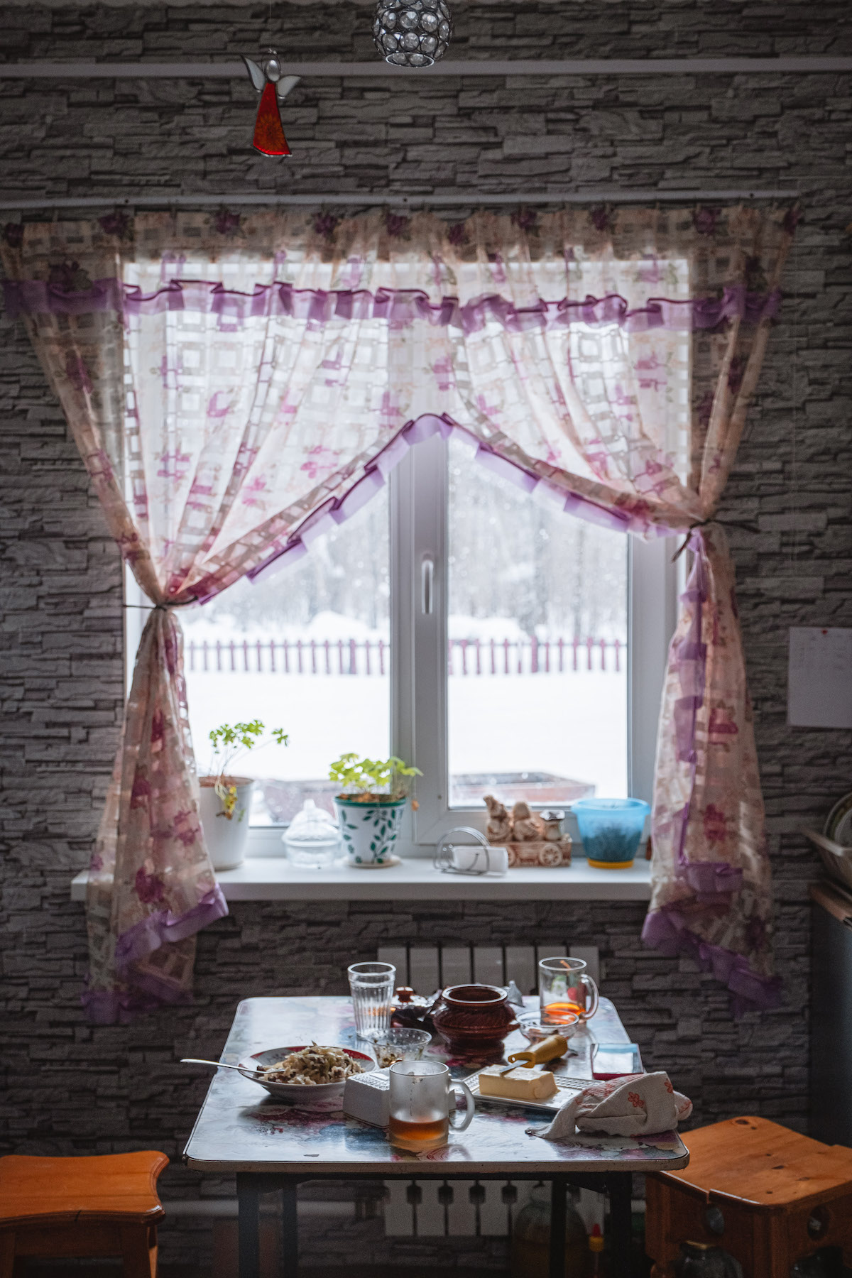 François Artusse, Udege people Russia – kitchen table with two stools, covered in mugs of tea, a dish of butter, a plate of food and different jars. In the background: a window framed by purple curtains.