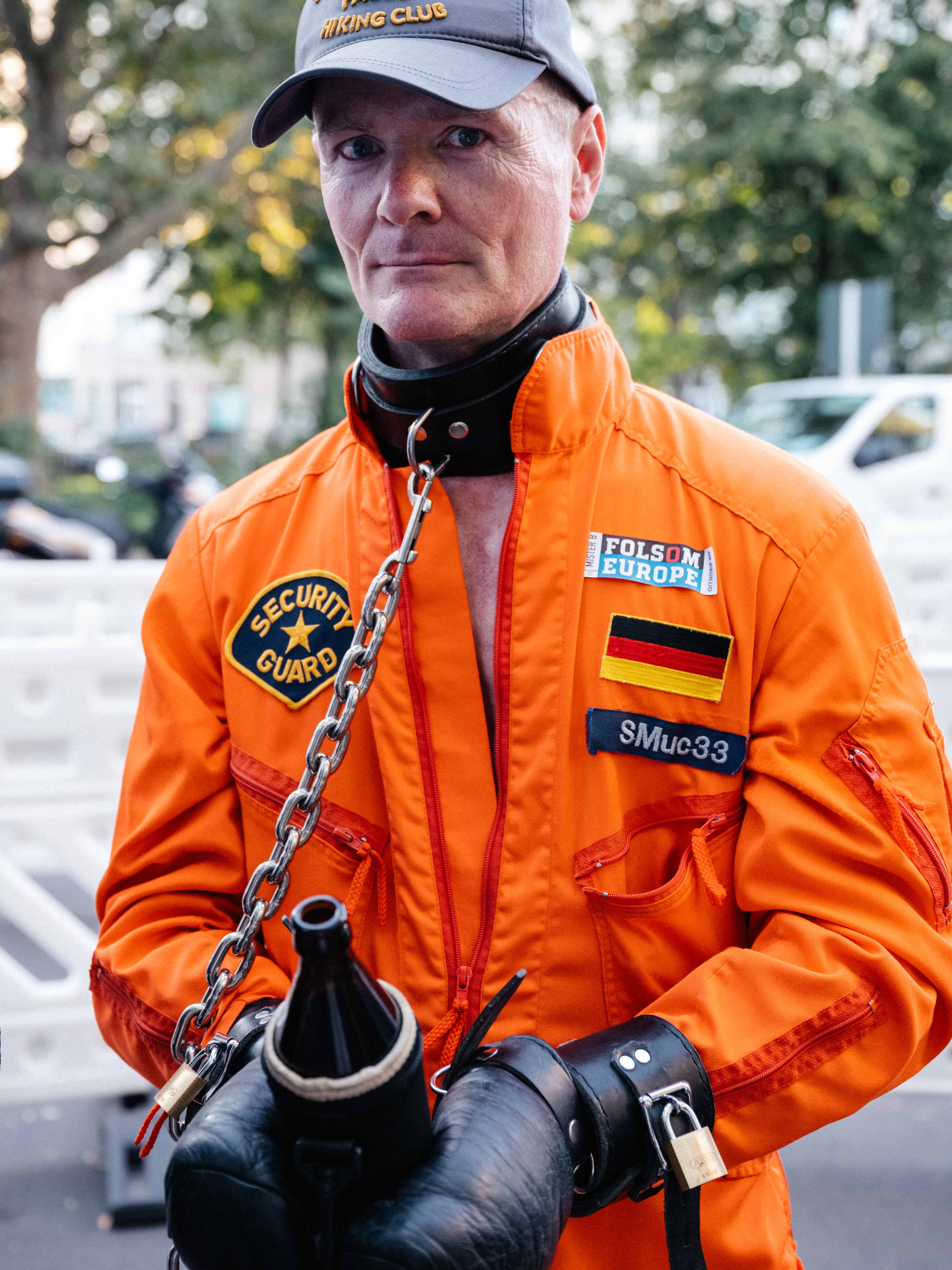 Close-up of a man wearing an orange jumpsuit, leather gloves, hand restraints and a choker.