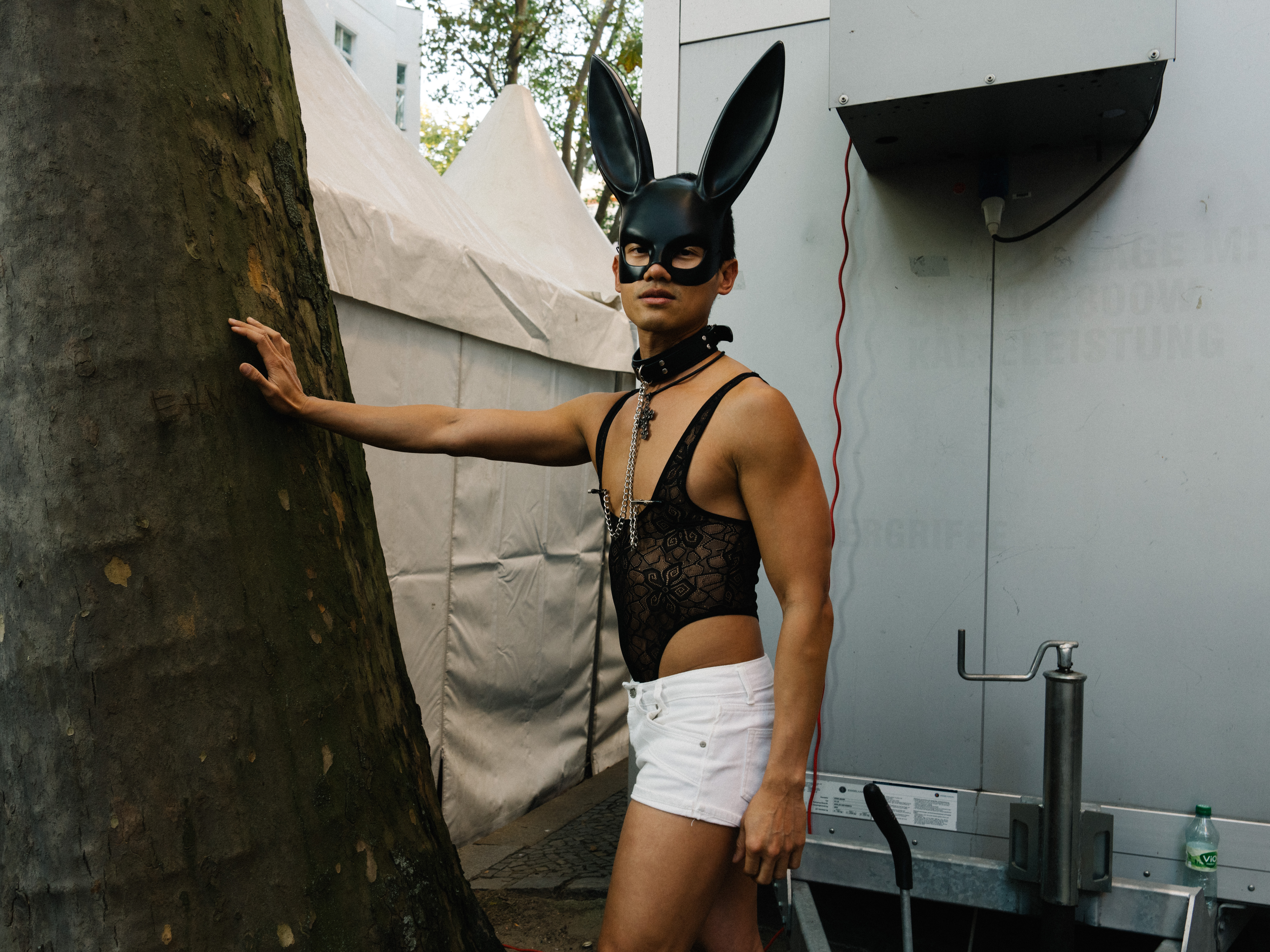 A person wearing white shorts, lace bodysuit, a choker with nipple chains, and a play bunny face mask.