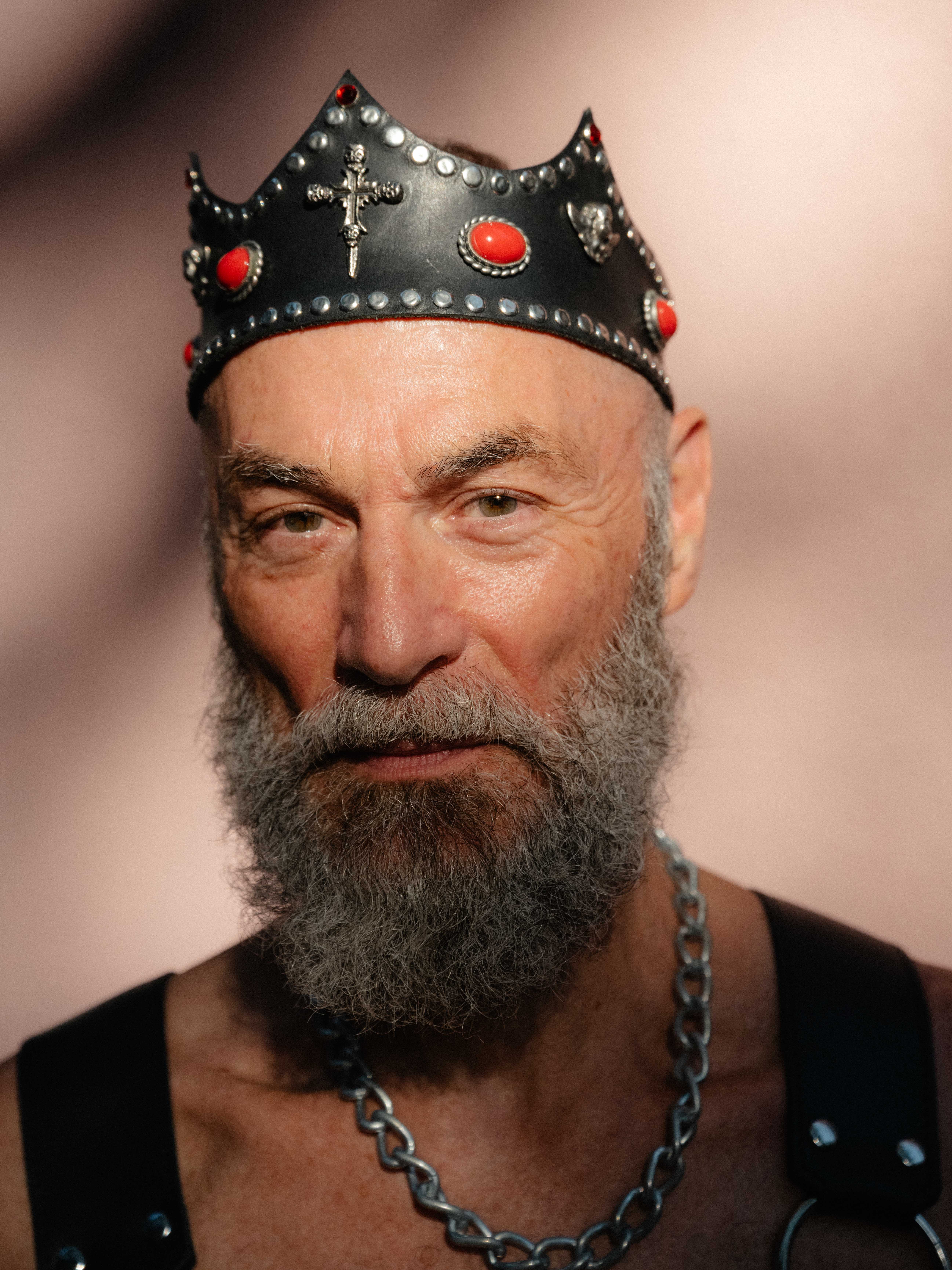 Close-up of a man with a thick grey beard wearing a leather studded crown, a harness and a thick chain around their neck.