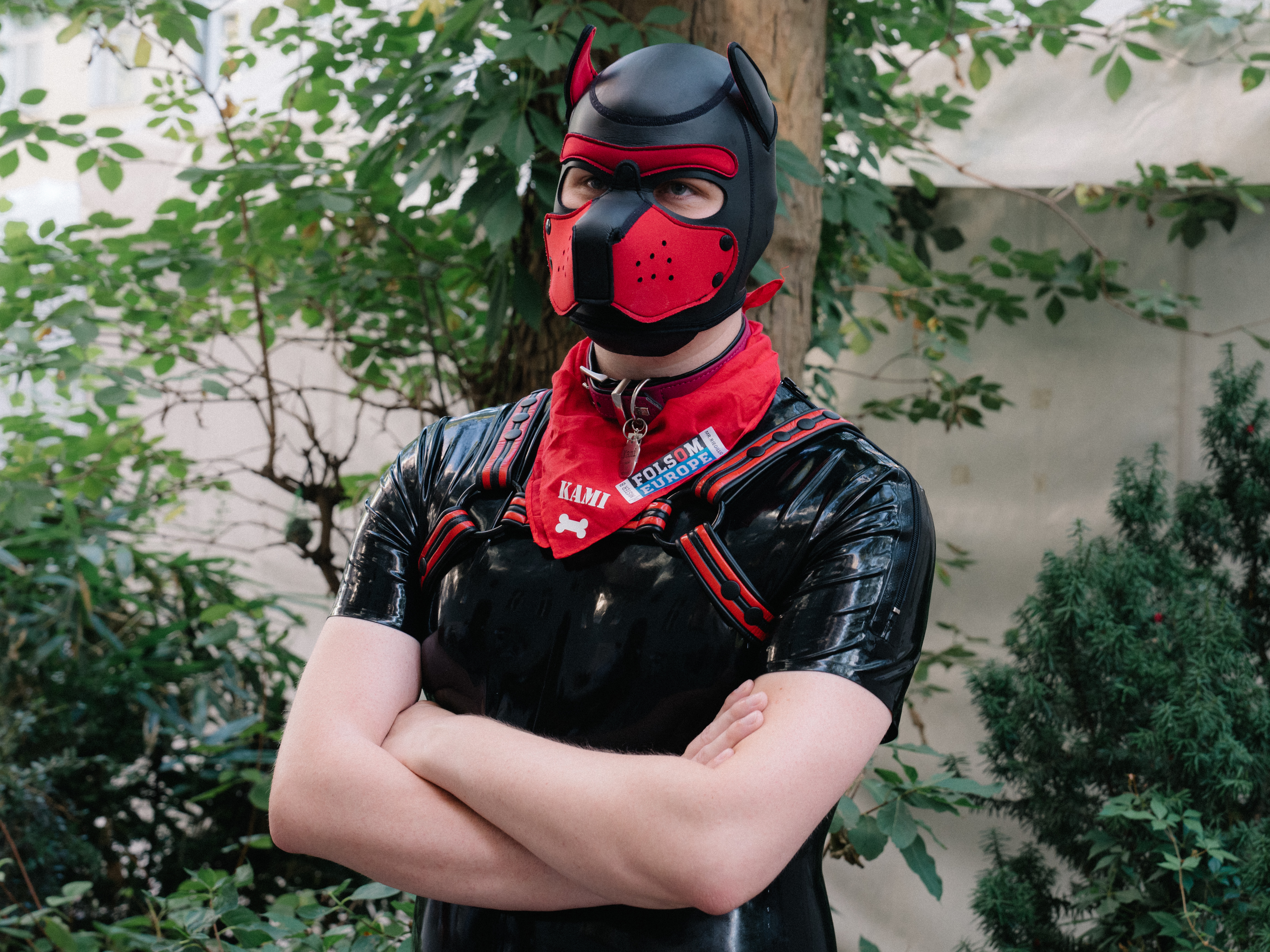 A person in short-sleeved black latex outfit, wearing a red and black mask, collar, bandana and harness; crossing their arms and looking off camera.