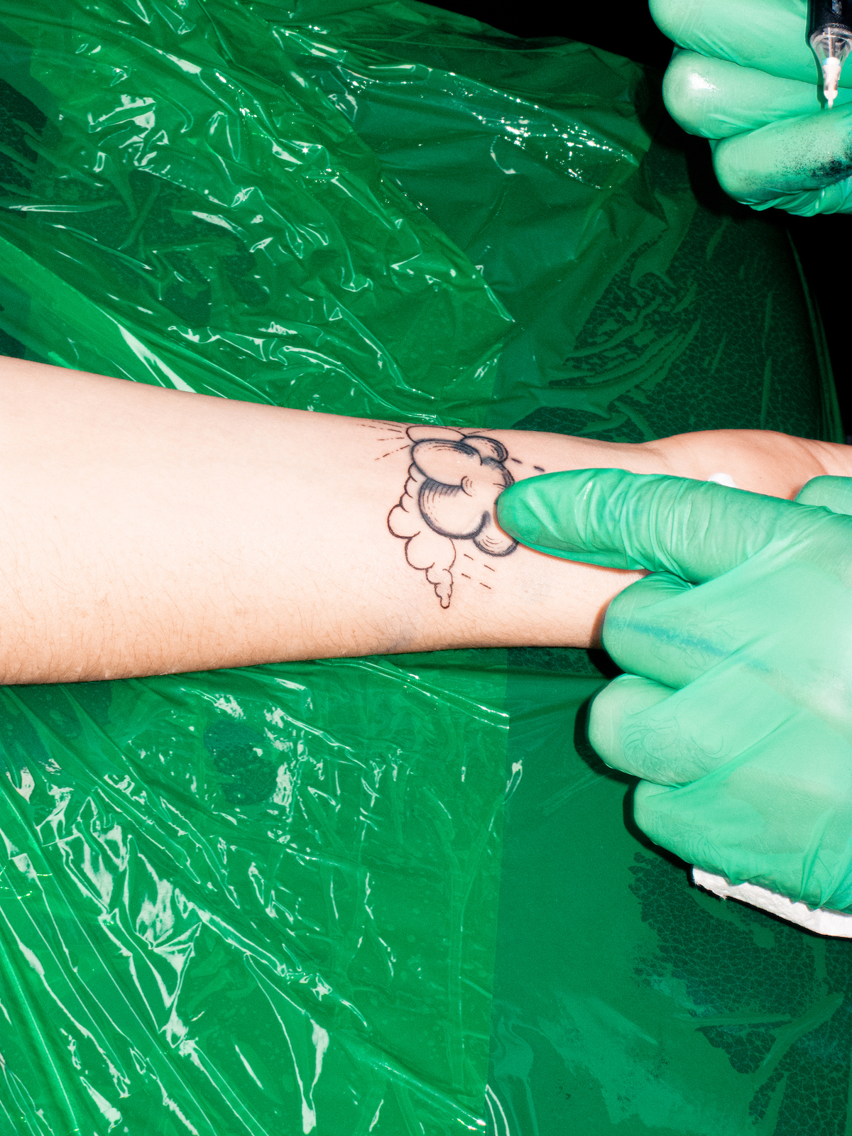 A photo of a person's wrist leaning on green plastic sheeting as it is tattooed by an artist's hands in green gloves.