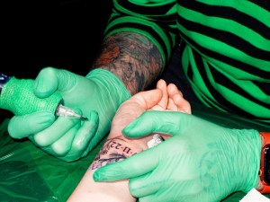 Foto van de handen van een tattoo artist met groene handschoenen die iemands pols tatoeëert