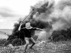 A man wearing a keffiyeh throwing a stone across a smoky field with a sling shot.