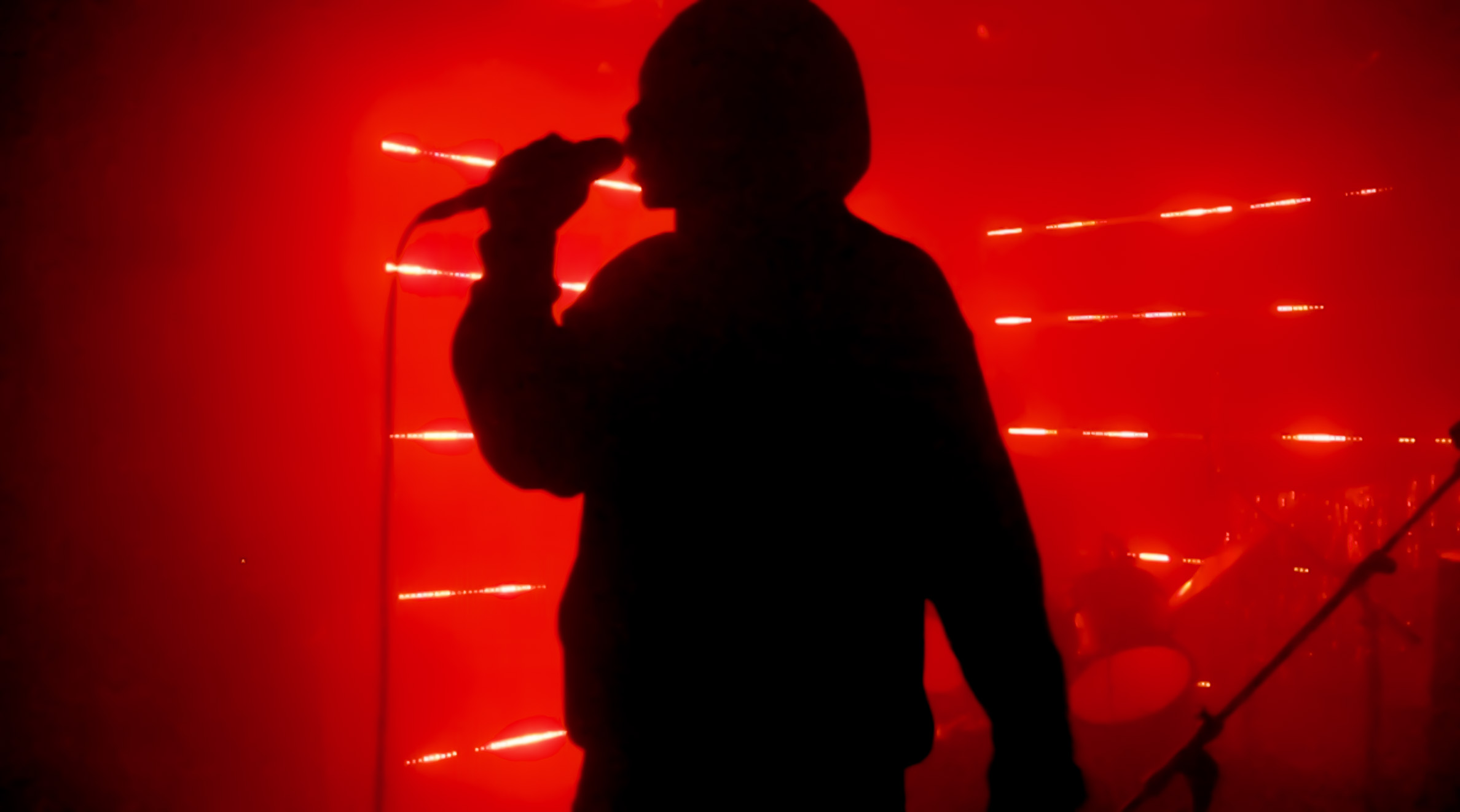 An image of bright red lights on the walls of a small stage, a man is singing into a microphone but he is just a black shadow from the lighting.