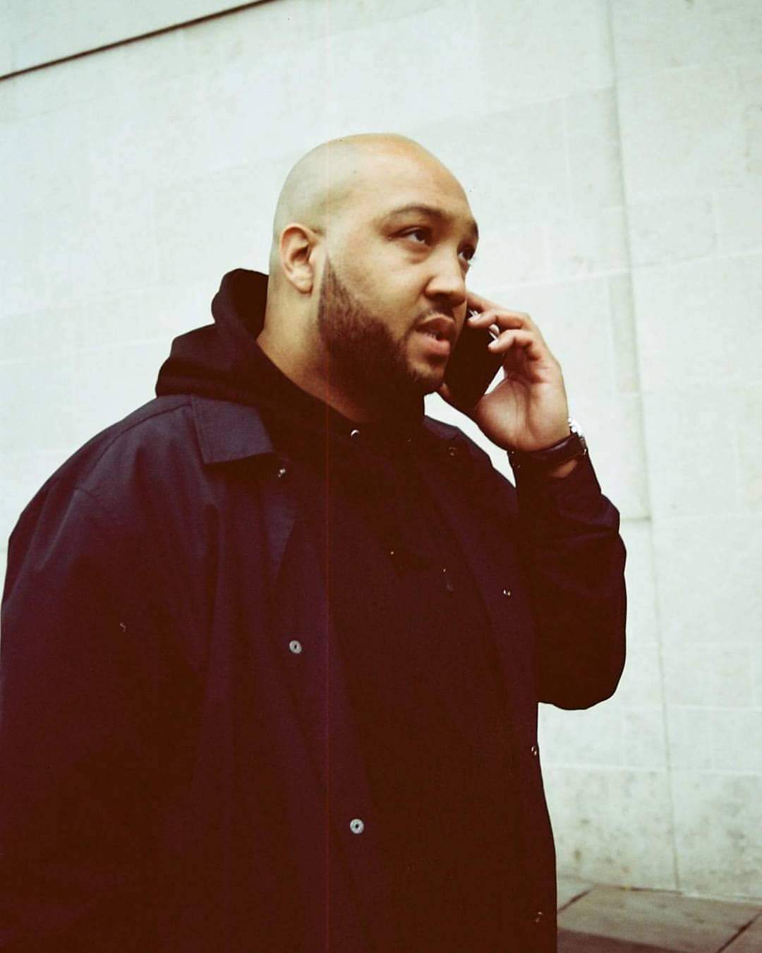 A photo of a black man with a bald head on the phone walking down the street. He's wearing a navy mac.