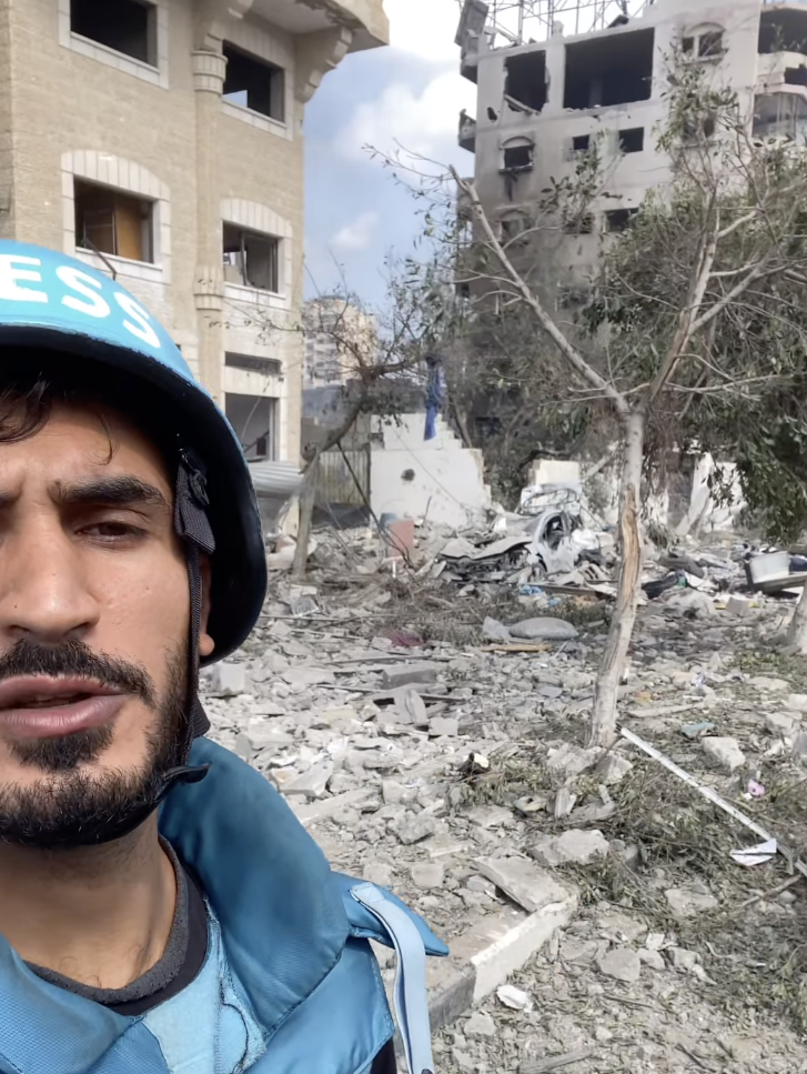 A man wearing a blue war journalist uniform takes a selfie in front of rubble