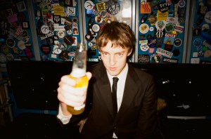 A young man in a suit nods towards the camera with a bottle of beer in his hand.