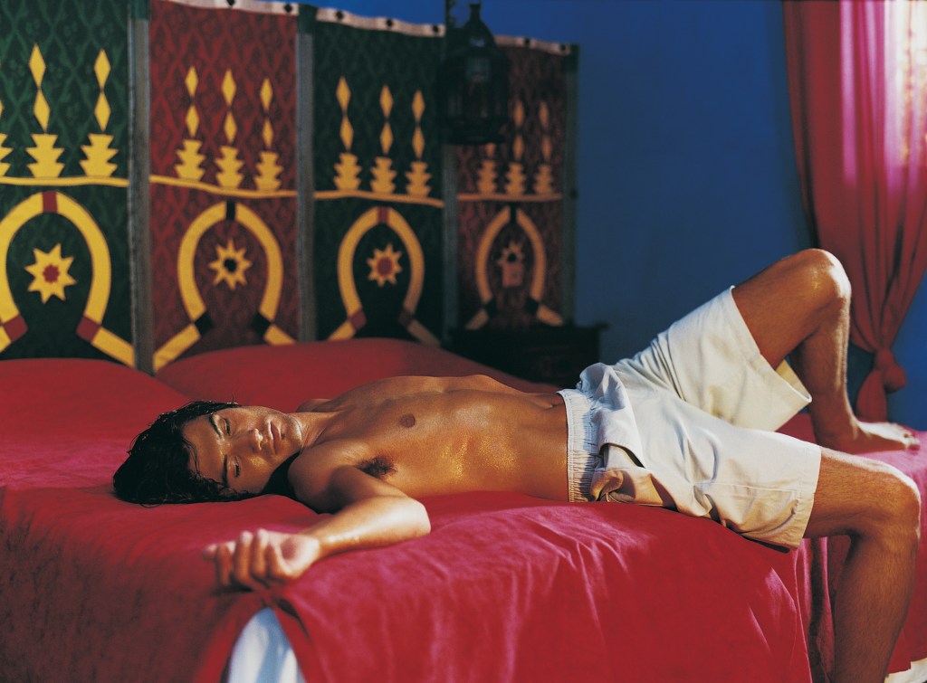 A sexy tanned man with abs lying on a bed of maroon sheets elegantly.