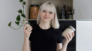 A photo of a woman with platinum blonde hair to the shoulders and bangs, holding jars in front of an organised cupboard and smiling to camera.