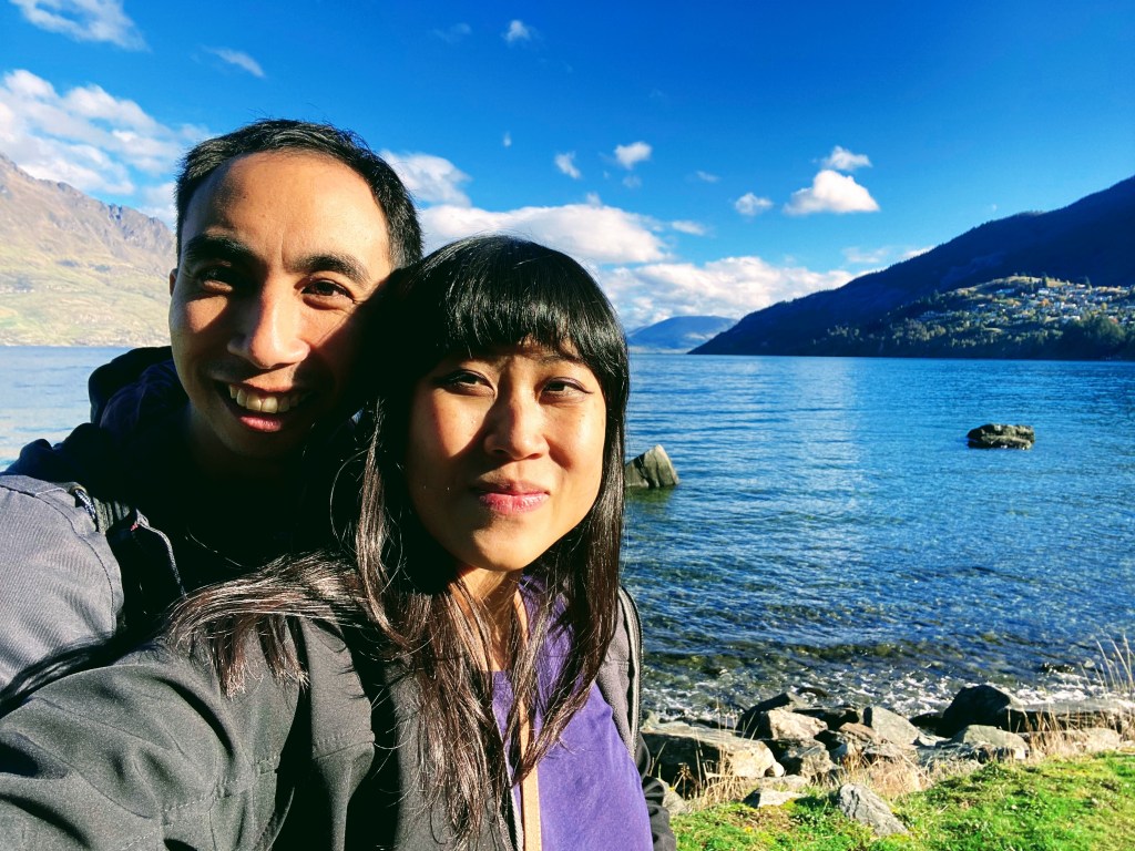 A couple smile while taking a photo together by a lake on a sunny day