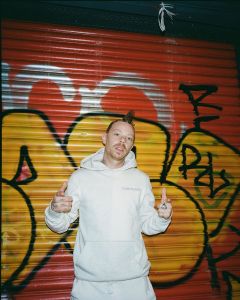 The dancehall musician MDotR wears a white hoody and poses in front of graffitied shutters