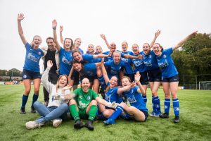 group photo of a women and queer football team on the pitch, they're wearing blue football jerseys except for one person in green and two people