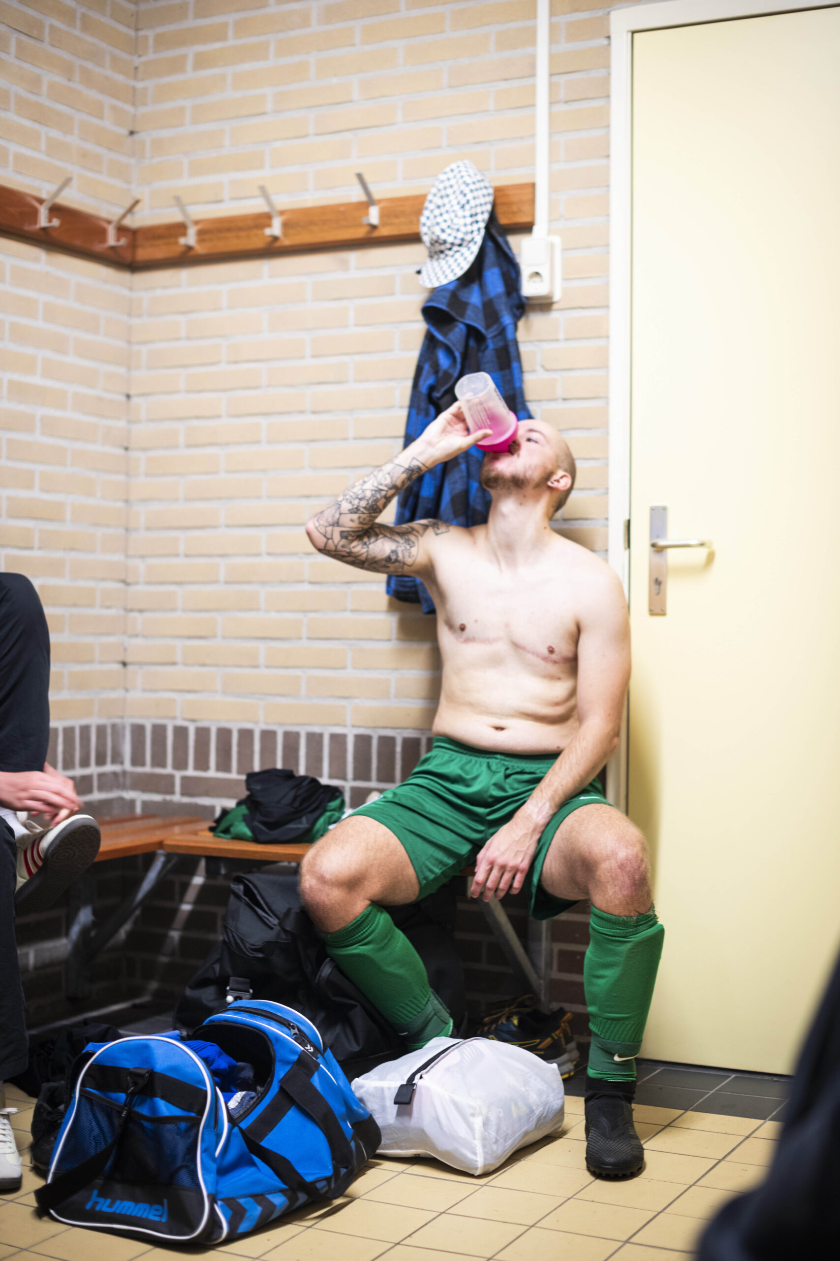 photo of a player drinking water and sitting on a bench in the locker room. They're wearing green shorts and high football socks and there are clothes and sports bags around them