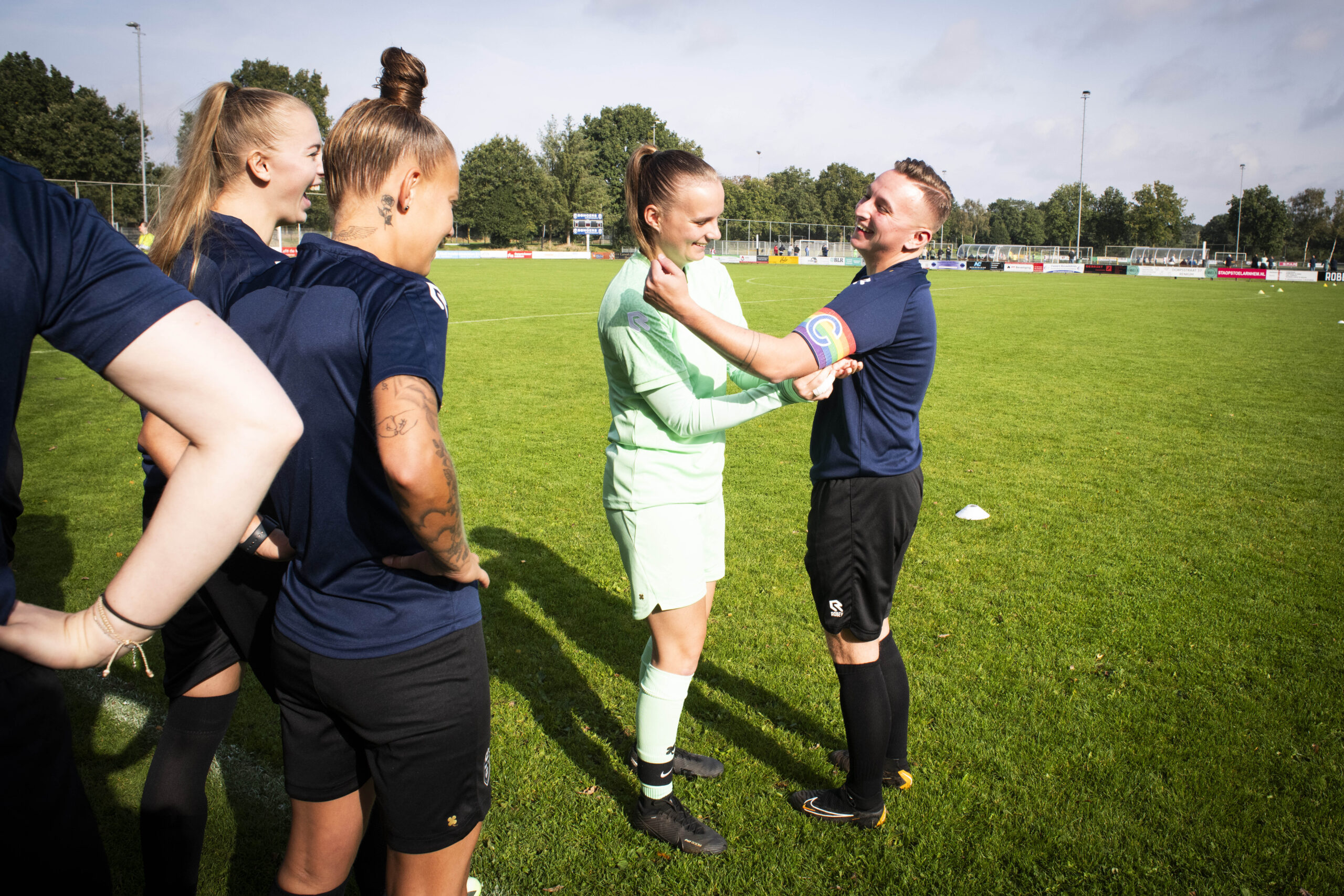 four players laughing on a football pitch, the goal keeper, dressed in light green, is putting a rainbow armband on another player's arm. The other three wear a dark blue jersey