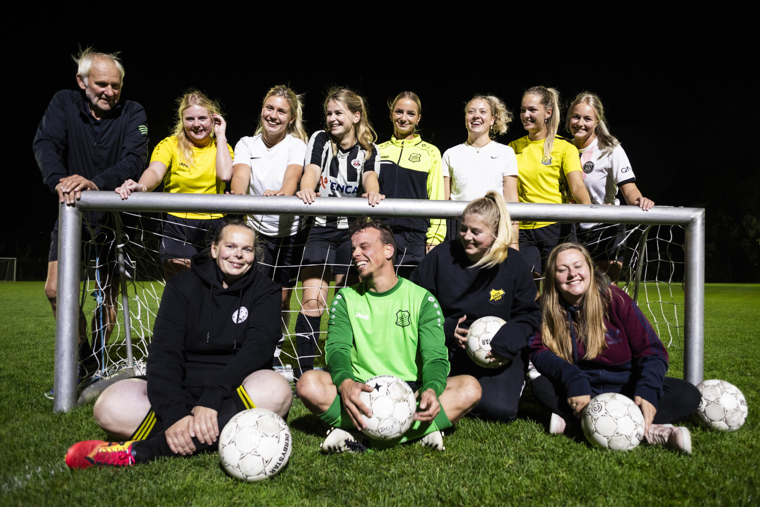 group photo of a women and queer football team near a small goal at night, they're wearing black, white and yellow football jerseys except for the goal keeper who's in green