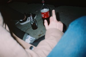 Flash-lit close-up of people sitting in the dark in front of a bottle of coke and a bottle of alcohol on the ground.