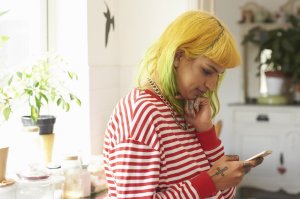 Unemployment: young woman with yellow hair, a white-and-red striped longsleeved shirt and tattoos, anxiously checking her phone in a white kitchen.