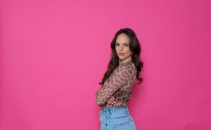 Gianna Bacio - woman with long wavy hair, wearing a mesh leopard print top and jeans and posing in front of a pink backdrop