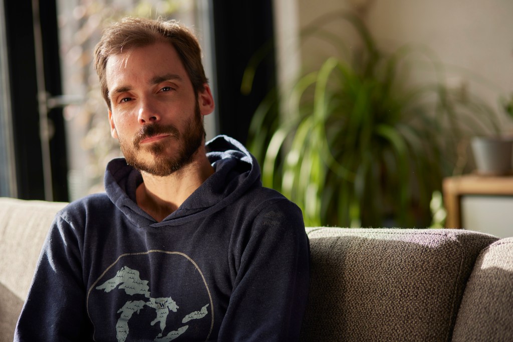 portrait of a white bearded man wearing a dark blue hoodie, he's sitting on his couch, looking at the camera. There's a plant in the background