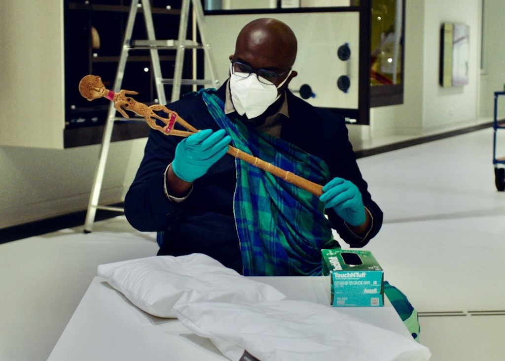 Colonial loot – Bald man wearing a face mask and disposable gloves, carefully examining a wooden object with the carving of a woman on top of it.