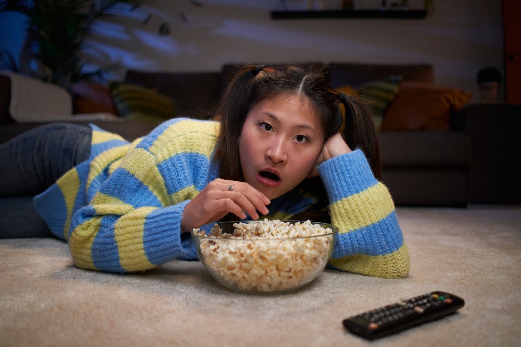 Point of view of a young Asian girl lying on the living room floor watching a movie at home, amazed by what's on TV, eating popcorn