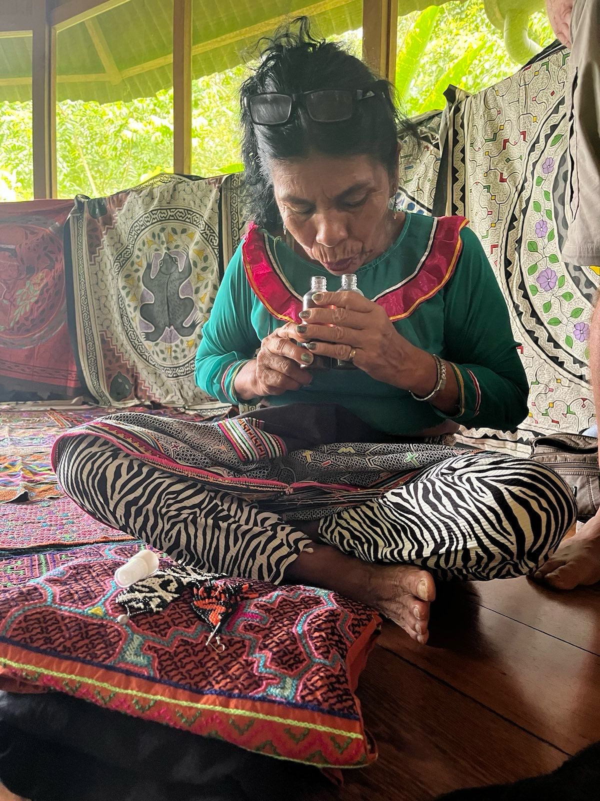 A woman sits cross-legged in a ritual space and blows into two small bottles.