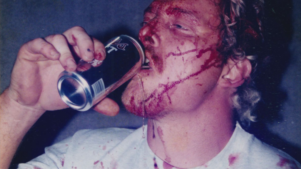 A photo of a white wrestler man with blood all over his face drinking a can of something.