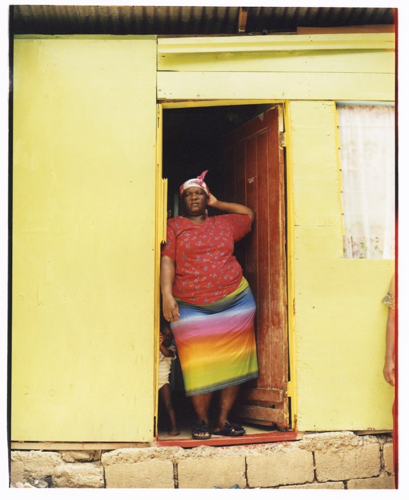 A young child hides behind an adult as they stand at the front door of their yellow house.