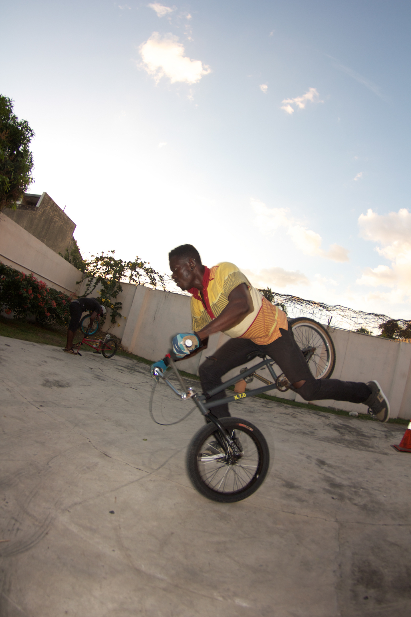 A man in a yellow polo shirt leans forward on one wheel on a BMX bike in a parking lot.