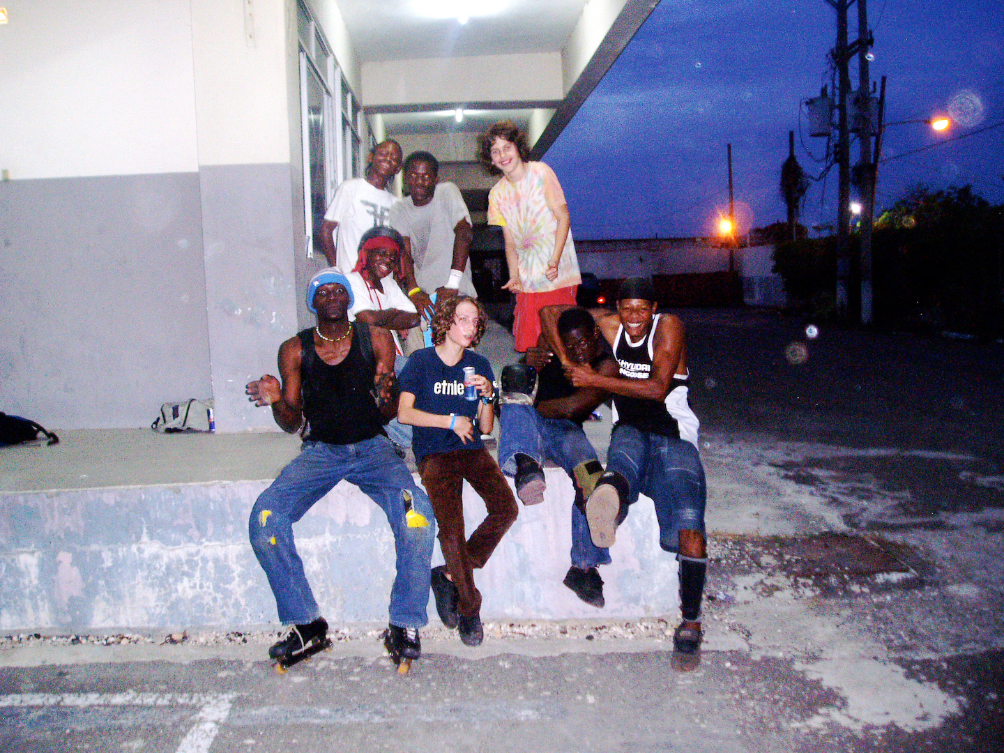 A group of BMX riders of varying ages sit together on a concrete kerb at night.