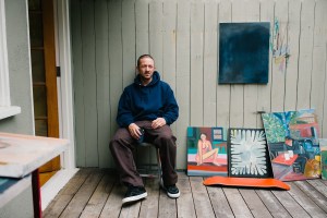 Skateboarder Mike Gigliotti sits on a chair next to three paintings and a skateboard