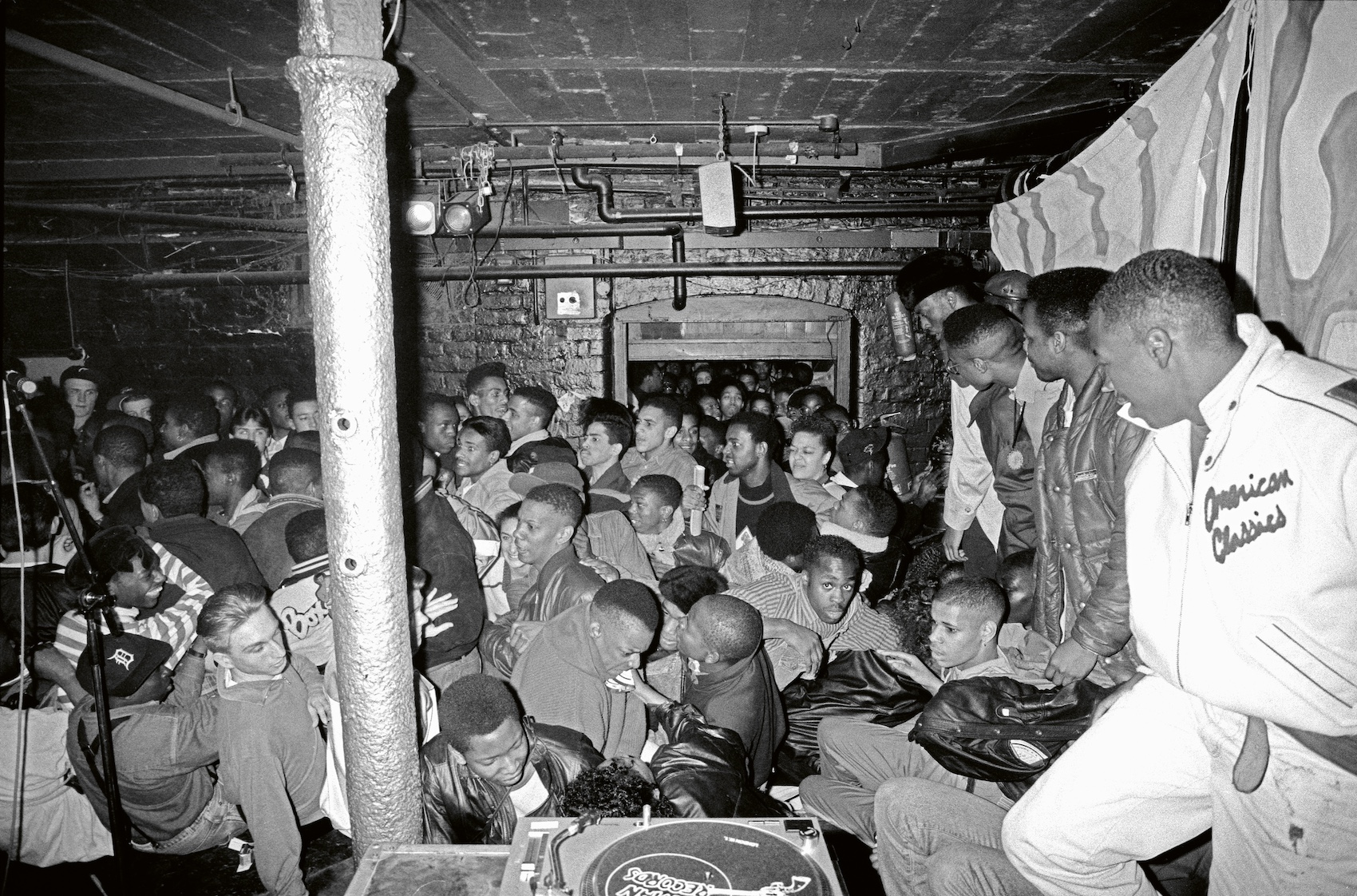 A small venue is packed with people and turntables can be seen in the foreground.