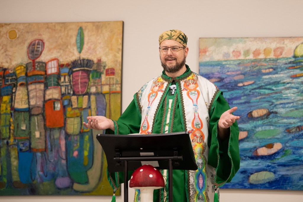 A religious leader in a green robe and weed-themed accessories stands at a mushroom pulpit.