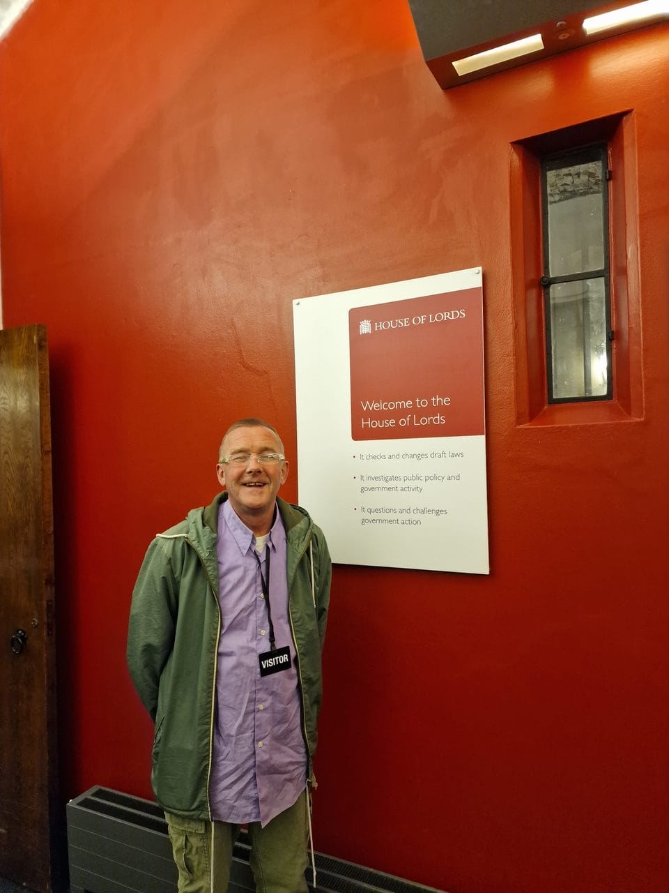 A man stands in a red corridor beside a sign that reads 'House of Lords'