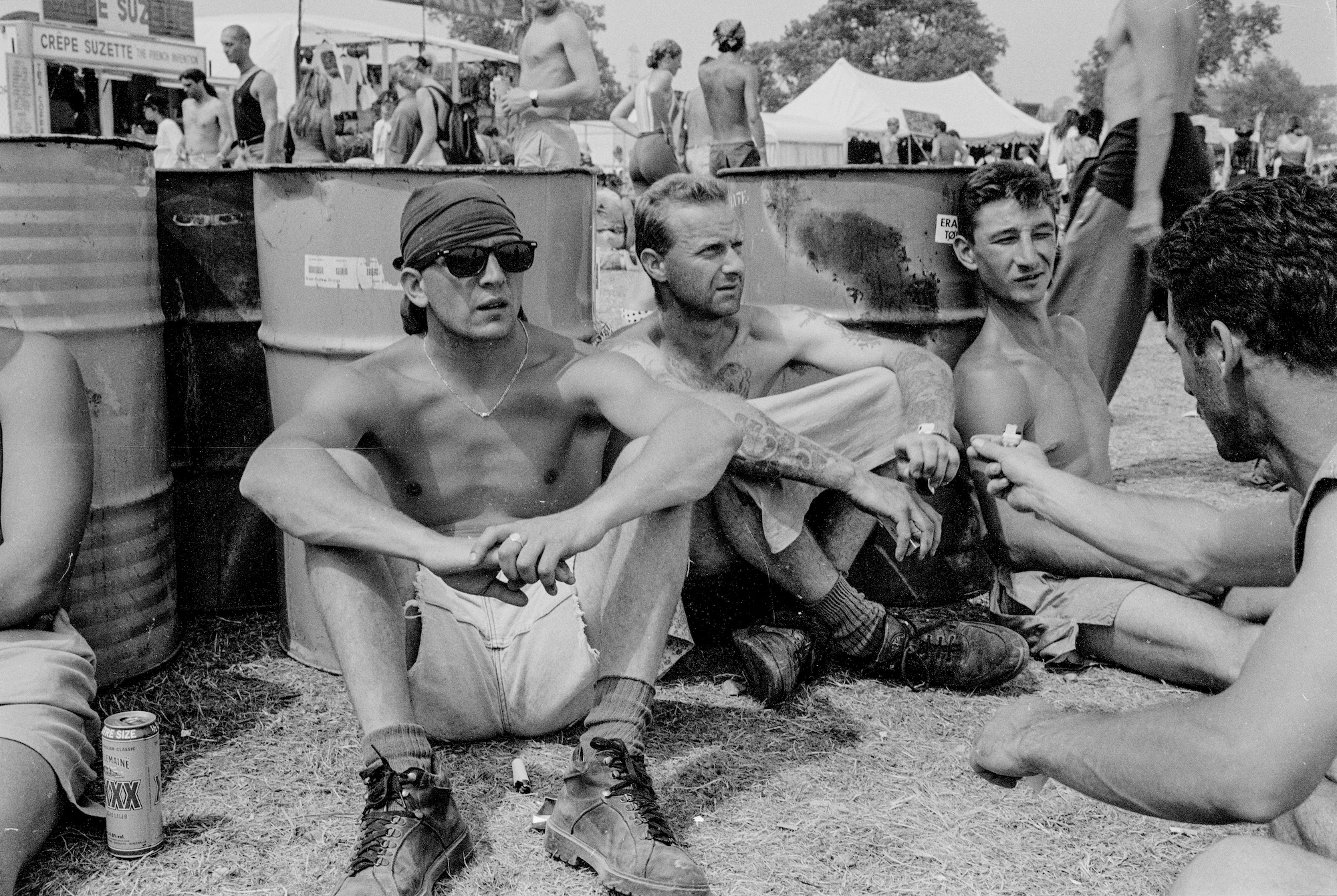 A group of men sit beside bins in a field and drink beer.